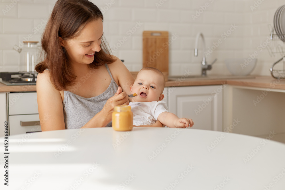 Good looking happy mother sitting with daughter or son in hands at the table and feeding baby with puree from the jar, indoor shot of mommy and eating child. Good looking happy mother sitting with daughter or son in hands at the table and feeding baby with puree from the jar, indoor shot of mommy and eating child.