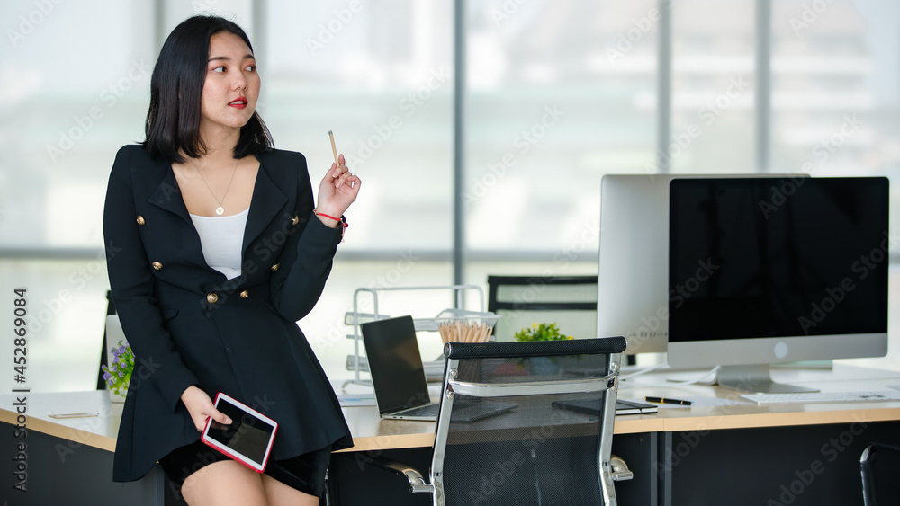 Young attractive Asian woman in black business suit sitting on table holding tablet with confidence in modern looking office with blurry windows background. Concept for modern office lifestyle