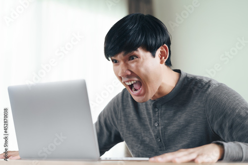 Angry shocked surprised Asian man looking at laptop computer screen sitting in the living room.