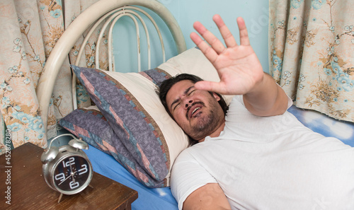 A tired, stubborn or lazy man refuses to get up from bed. Gesturing to stop waking him up.