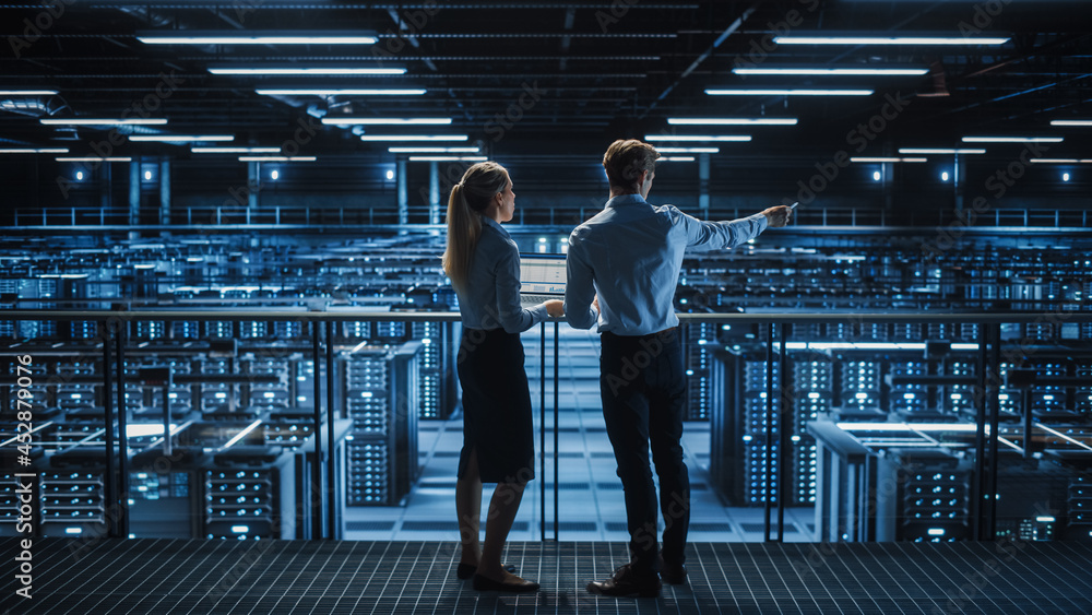 Data Center Male It Specialist and Female System administrator Talk, Use Laptop. Server Farm Cloud Computing Facility with Two Information Technology Professionals working on Cyber Secury. Data Center Male It Specialist and Female System administrator Talk, Use Laptop. Server Farm Cloud Computing Facility with Two Information Technology Professionals working on Cyber Secury.
