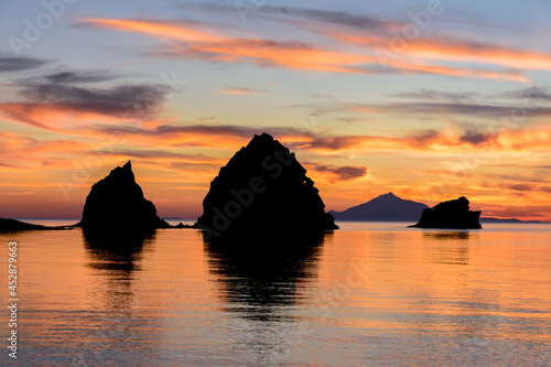 Three rocky islets against the backdrop of an island at sunset with orange and cloudy skies.