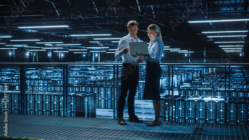 Data Center Female e-Business Enrepreneur and Male IT Specialist talk, Use Laptop. Two Information Technology Engineers on Bridge Overlooking Big Cloud Computing Server Farm.