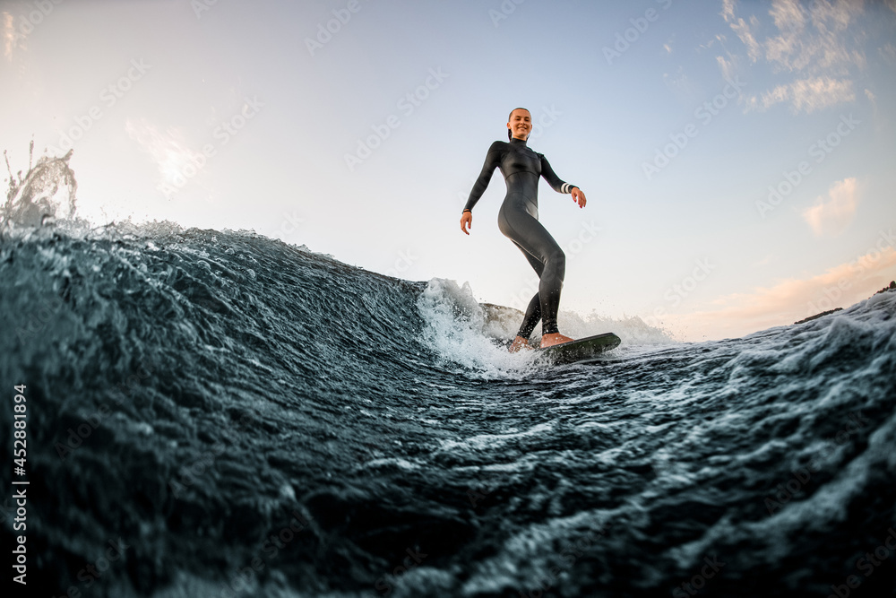 Young handsome woman wakesurfing on the board down the river against the background of blue sky
