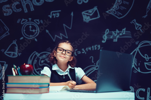 little schoolgirl in glasses studying with computer, school blackboard background, back to school