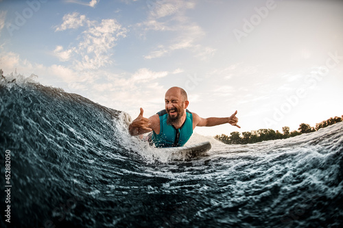 cheerful male wakesurfer lying on board rides down wave and show hands gestures and winks