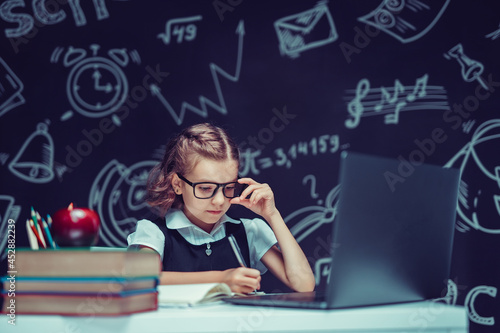little schoolgirl in glasses studying with computer, school blackboard background, back to school