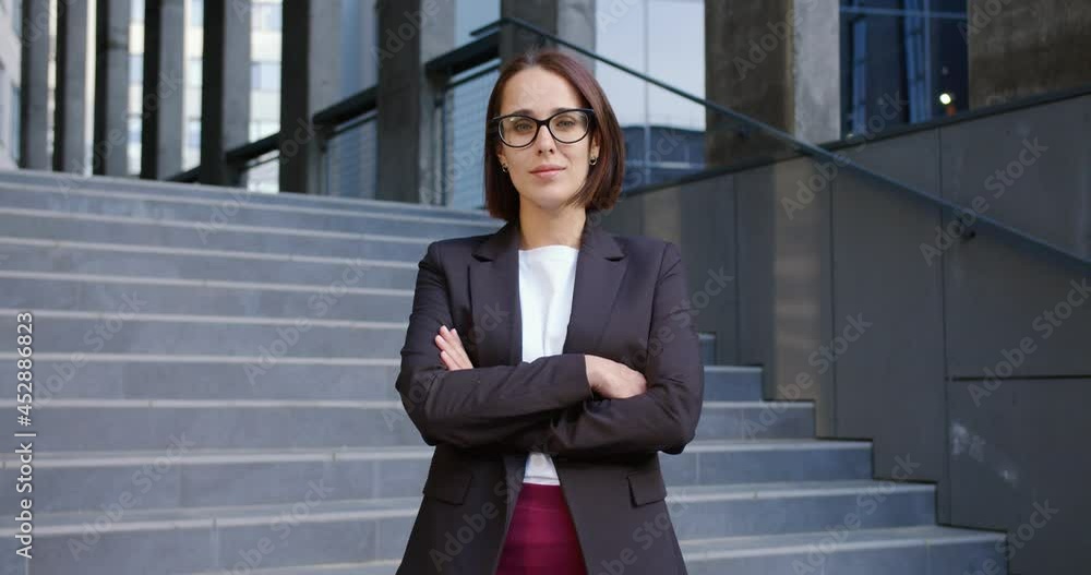 Portrait of pretty young businesswoman looking at camera while standing outdoors in city. Caucasian confident female financial consultant stands outside on stairs near office center. Business style Portrait of pretty young businesswoman looking at camera while standing outdoors in city. Caucasian confident female financial consultant stands outside on stairs near office center. Business style