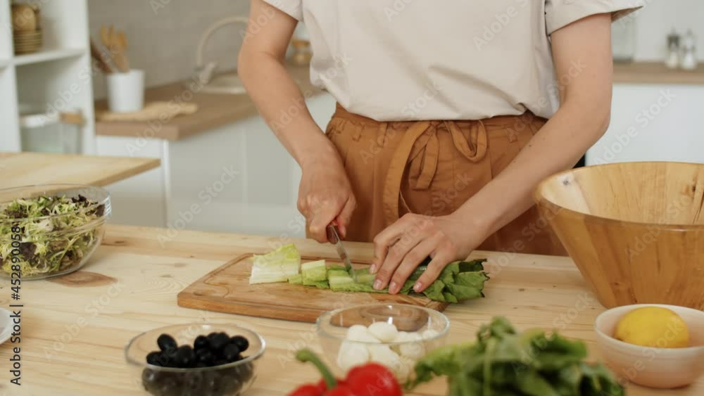 Tilt down shot of woman cutting fresh lettuce and putting leaves into wooden salad bowl while preparing meal in kitchen at home Tilt down shot of woman cutting fresh lettuce and putting leaves into wooden salad bowl while preparing meal in kitchen at home