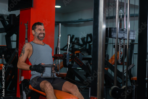 Fitness training in sport gym of active male with beard sitting and pulling heavy weight on exercise equipment