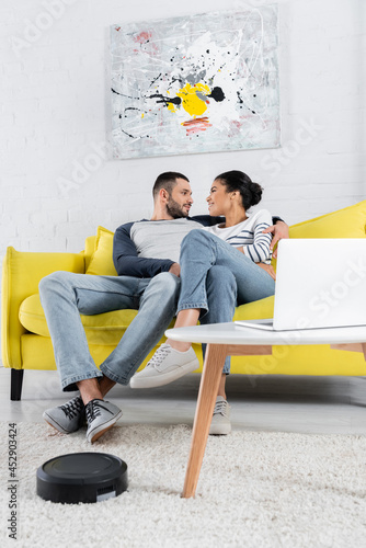 Side view of man hugging african american girlfriend near blurred laptop and robotic vacuum cleaner