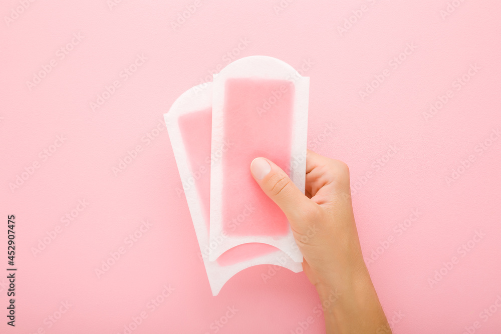 Young adult woman hand holding two wax strips on light pink table background. Pastel color. Closeup. Female product for smooth body skin. Top down view. Young adult woman hand holding two wax strips on light pink table background. Pastel color. Closeup. Female product for smooth body skin. Top down view.