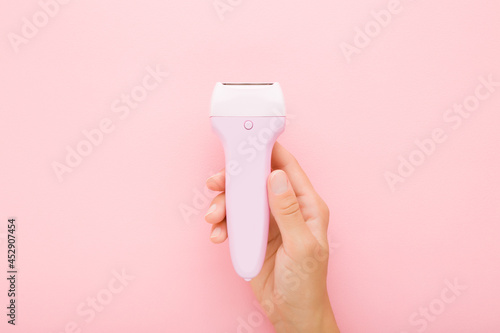 Young adult woman hand holding electric epilator on light pink table background. Pastel color. Closeup. Female product for smooth body skin. Top down view.