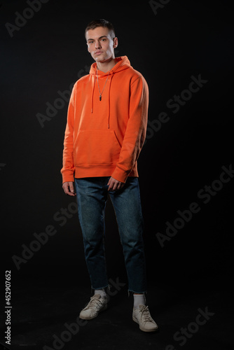Young handsome man in jeans and orange hoodie posing while standing on a black background.