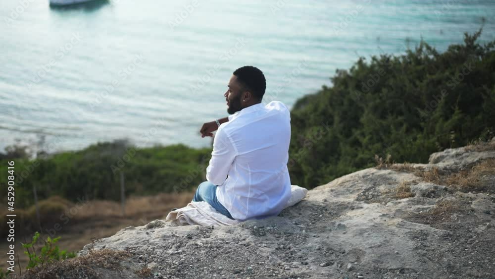 Back view of relaxed African American LGBT man sitting on cliff admiring beauty of blue Mediterranean sea. Happy carefree gay tourist resting on vacations. Tranquility and freedom concept Back view of relaxed African American LGBT man sitting on cliff admiring beauty of blue Mediterranean sea. Happy carefree gay tourist resting on vacations. Tranquility and freedom concept