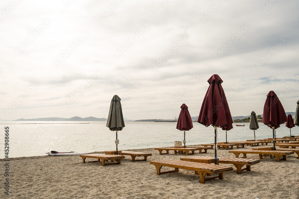 Naklejka premium Empty wooden deck beds and folded umbrellas on sand near Aegean Sea. Lounge chill zone on city beach in Athens, Greece