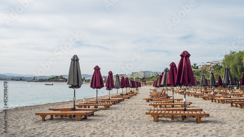 Fototapeta Naklejka Na Ścianę i Meble -  Empty wooden deck beds and folded umbrellas on sand near Aegean Sea. Lounge chill zone on city beach in Athens, Greece