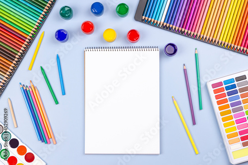 Creative art work supplies background. Watercolor, brushes, pencils, open blank notebook on light gray table. Top view, flat lay