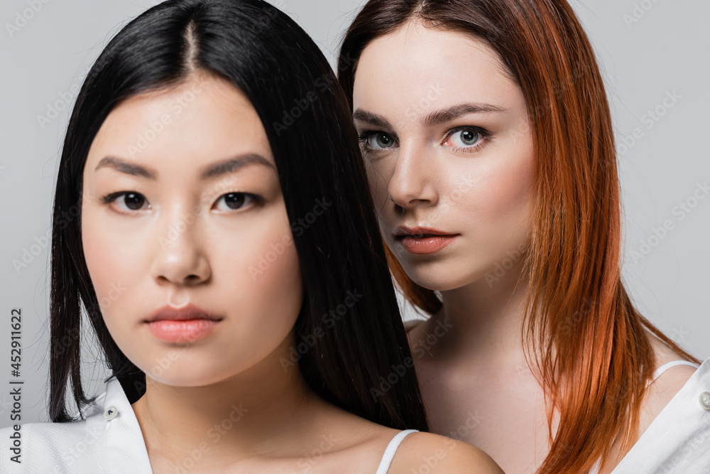 redhead woman looking at camera near blurred asian model isolated on grey redhead woman looking at camera near blurred asian model isolated on grey