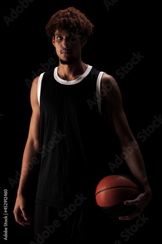 Professional basketball player holding a ball against black background. Serious concentrated african american man in sports uniform © Nikola Spasenoski