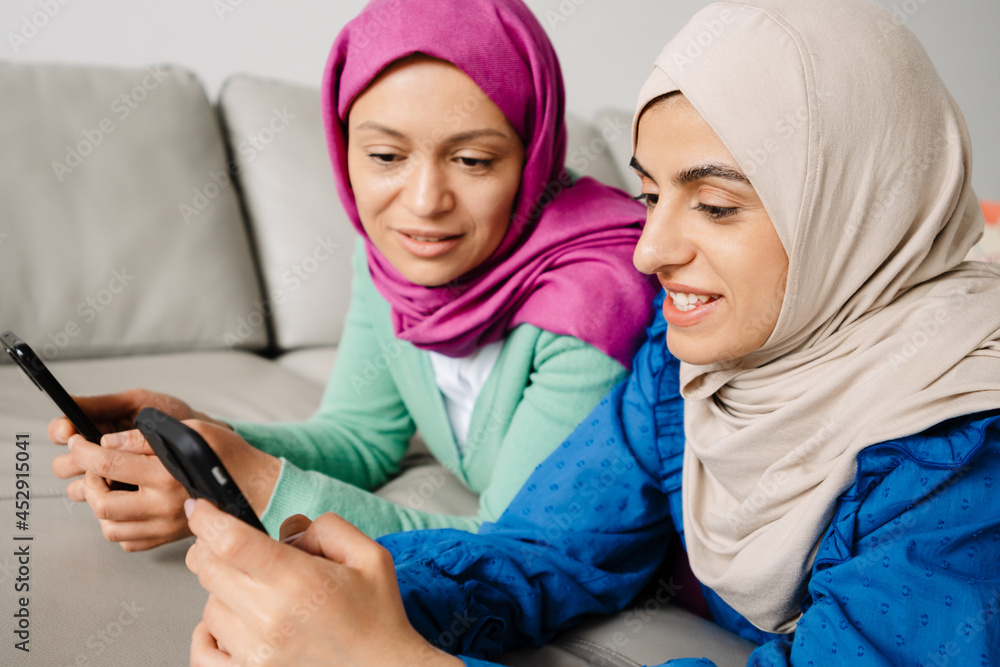 Fototapeta premium Two women in headscarf using mobile phones while resting on sofa