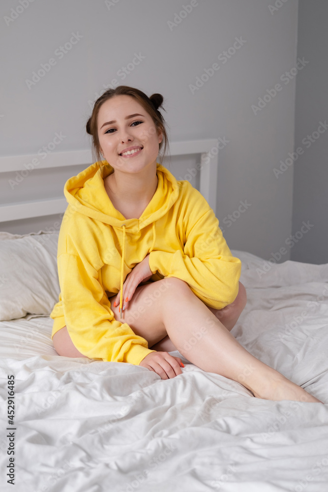 happy cheerful brunette girl in yellow hoodie in white bedroom sitting on bed with white bed linens. happy cheerful brunette girl in yellow hoodie in white bedroom sitting on bed with white bed linens.