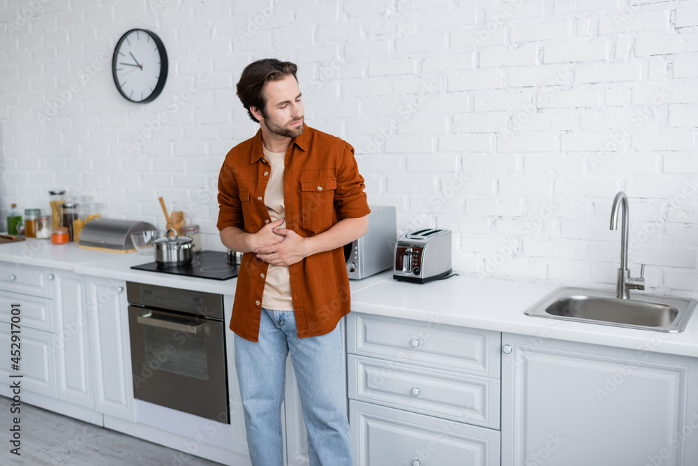 man suffering from stomach pain while standing with closed eyes in kitchen man suffering from stomach pain while standing with closed eyes in kitchen