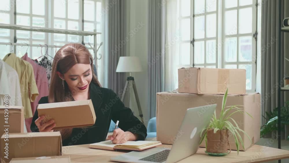 Young Asian Shemale Business Owner With Boxes On The Table Happy Online Sales Job, Use The Laptop, Get An Order From Customers, Take Notes, And Make Arrangements For Delivery By Post vídeo do Stock | Adobe Stock