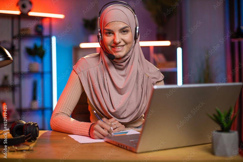 Studying online, remote work, freelance concept. Beautiful high-skilled modern muslim woman in hijab and headphones, sitting at the table at home with laptop and making notes