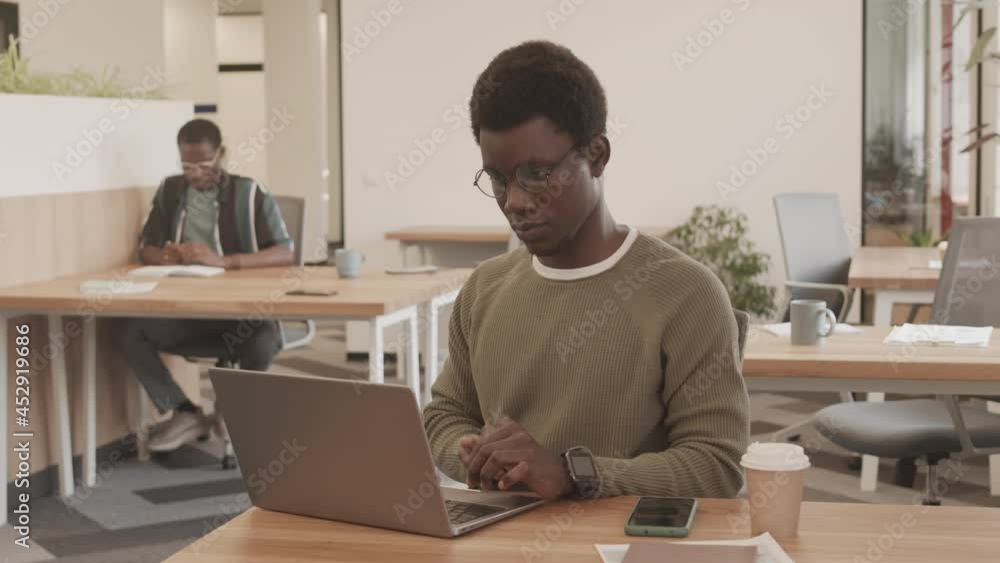 Medium shot of young African-American man in glasses and smart casualwear working on laptop sitting by desk in modern office Medium shot of young African-American man in glasses and smart casualwear working on laptop sitting by desk in modern office
