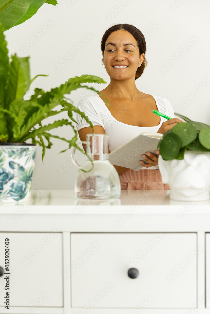 Thoughtful ethnic woman with notebook between assorted plants at home Thoughtful ethnic woman with notebook between assorted plants at home
