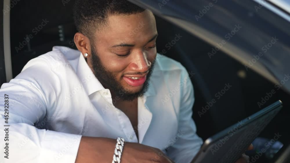 Smiling gay man reading messages in social media using app on tablet sitting in car. Portrait of happy African American confident handsome gay messaging online in sunset sunshine Smiling gay man reading messages in social media using app on tablet sitting in car. Portrait of happy African American confident handsome gay messaging online in sunset sunshine