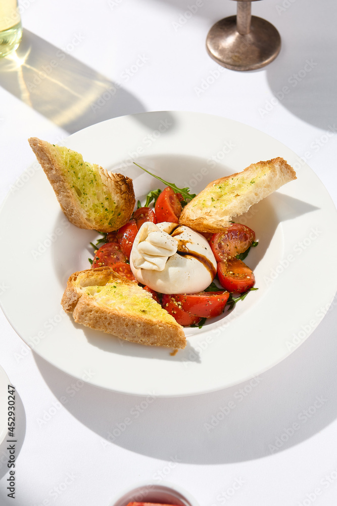 Traditional Italian salad with Burrata cheese and tomatoes. Fancy caprese with burrata cheese, cherry tomatoes, pesto and balsamic sauce. Summer dish with mozzarella cheese on white table with shadow. Traditional Italian salad with Burrata cheese and tomatoes. Fancy caprese with burrata cheese, cherry tomatoes, pesto and balsamic sauce. Summer dish with mozzarella cheese on white table with shadow.
