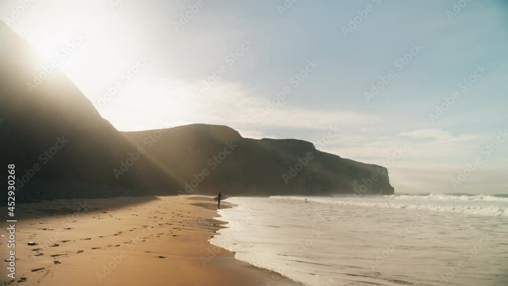 Silhouette of lonely surfer in wetsuit stand on edge of surf waves on epic beach. Beautiful inspiring background of calm majestic landscape. Surfing vibes lifestyle. Sunrise at beach.Portugese winter Silhouette of lonely surfer in wetsuit stand on edge of surf waves on epic beach. Beautiful inspiring background of calm majestic landscape. Surfing vibes lifestyle. Sunrise at beach.Portugese winter