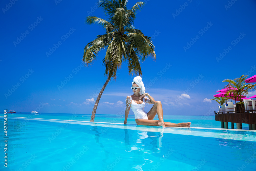 Elegant sexy woman in the white bikini on the sun-tanned slim and shapely body is posing near the swimming pool on Maldives. Perfect body bikini model in luxury resort on Maldives. Wellness.