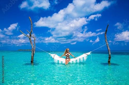 Elegant sexy woman in the white bikini on the sun-tanned slim and shapely body is posing on hammock in ocean on Maldives. Perfect body bikini model in luxury resort on Maldives. Luxury travel.