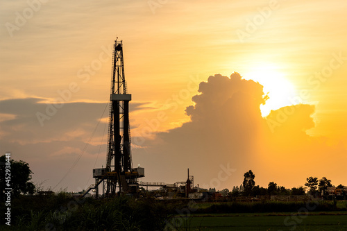 Fotografi Silhouette black shadow of oil exploration drilling rig structure among the orange sunrise sky and agriculture field environemnt