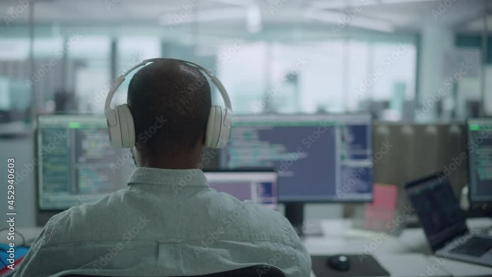 Office: Professional Black IT Programmer Uses Headphones while Working on Desktop Computer. Male Website Developer, Software Engineer Developing App, Video Game. Listening to Podcast, Music. Back View Office: Professional Black IT Programmer Uses Headphones while Working on Desktop Computer. Male Website Developer, Software Engineer Developing App, Video Game. Listening to Podcast, Music. Back View