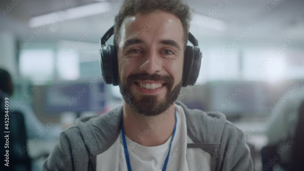 Office: Portrait of Happy IT Programmer Wearing Headphones Working on Desktop Computer, Looking at Camera and Smiling. Male Software Engineer Developing App, Video Game. Listening to Podcast, Music Office: Portrait of Happy IT Programmer Wearing Headphones Working on Desktop Computer, Looking at Camera and Smiling. Male Software Engineer Developing App, Video Game. Listening to Podcast, Music