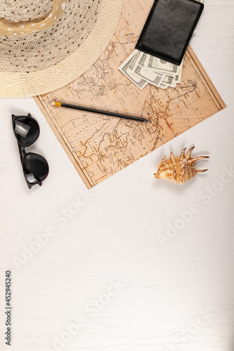 world map, straw hat, black leather wallet with money, sunglasses, pencil and shell on a white wooden table