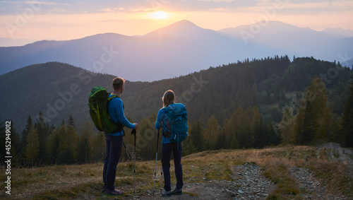 Rear view of lover pair of hikers standing on mountain trail and leaning on trekking sticks watching the mountain scenes in summer evening. Concept of travelling, hiking and active leisure. © anatoliy_gleb