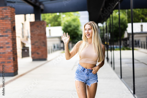 Hi there. Beautiful young woman in hat and sunglasses waving top someone and smiling while walking by the street