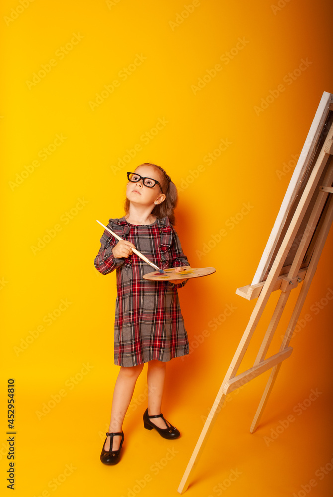 Portrait of a beautiful child girl holding a wooden palette and paint brush against a yellow wall. Smiling child with an artistic palette and an artist's brush in their hands. Portrait of a beautiful child girl holding a wooden palette and paint brush against a yellow wall. Smiling child with an artistic palette and an artist's brush in their hands.