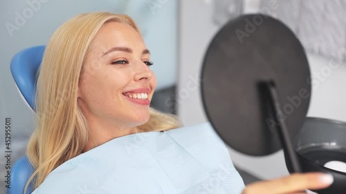Positive smiling satisfied young woman patient hold look in the mirror sitting in chair in dental clinic light office medical center with modern tools equipment. Healthy lifestyle treatment concept