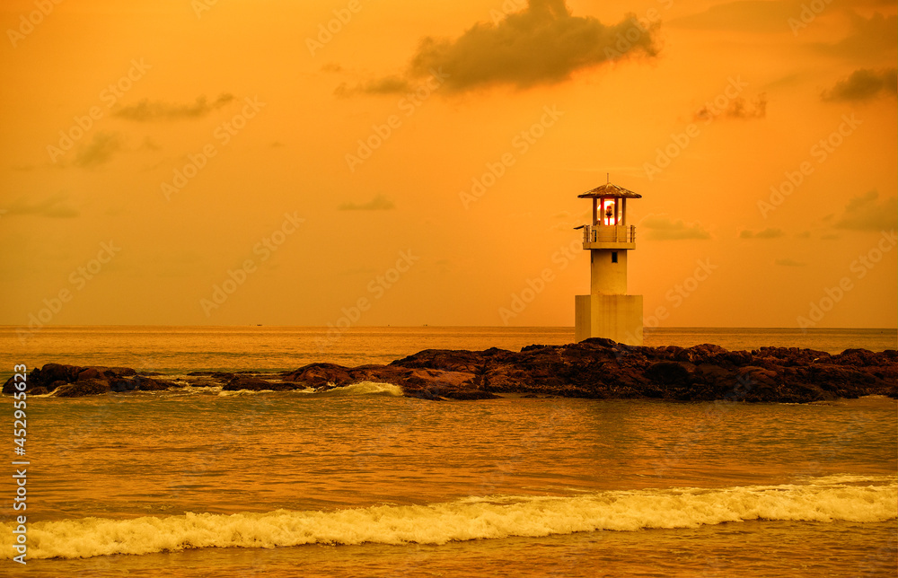 Panorama view of lighthouse on small rock island with orange sky and dramatic clouds at sunset. Taken in south of Thailand