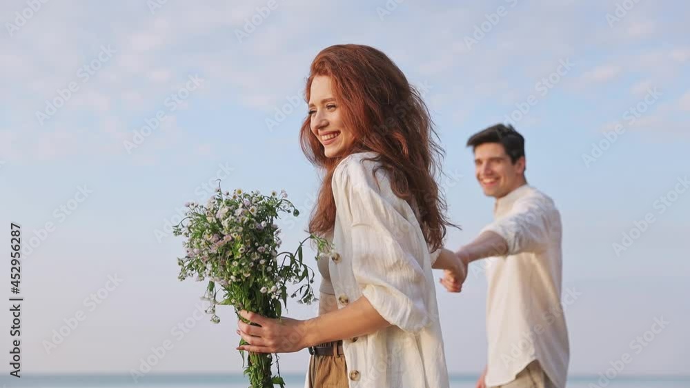 Happy young couple two friends family 20s in white clothes woman hold flowers bouquet hold man hand leading him somewhere at sunrise over sea beach ocean outdoor seaside in summer day sunset evening Happy young couple two friends family 20s in white clothes woman hold flowers bouquet hold man hand leading him somewhere at sunrise over sea beach ocean outdoor seaside in summer day sunset evening