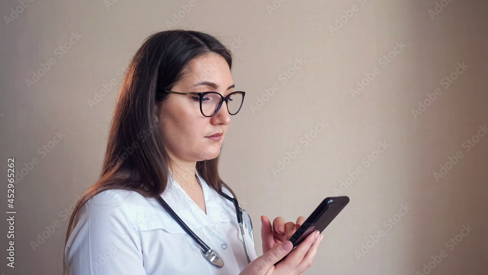 Pretty young female doctor with glasses and stethoscope texts message on mobile phone standing near beige wall in hospital office closeup Pretty young female doctor with glasses and stethoscope texts message on mobile phone standing near beige wall in hospital office closeup