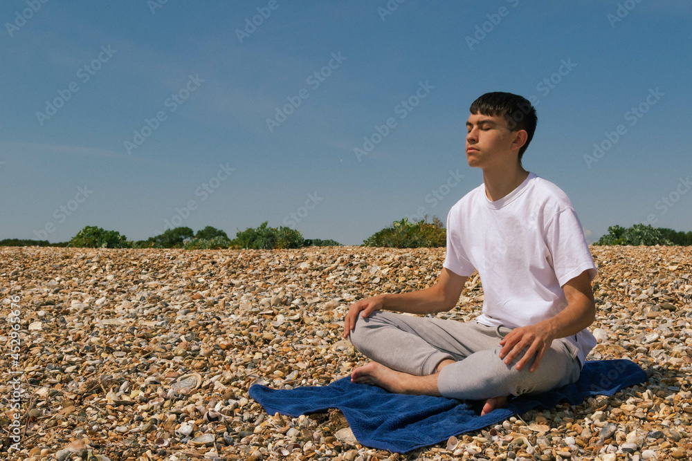 A Caucasian teenage boy meditating on a stony beach with his hands resting on his knees