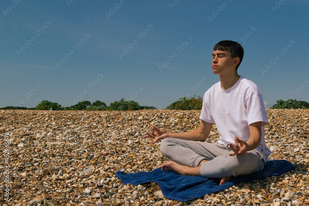 A Caucasian teenage boy meditating on a stony beach with his hands in the Shuni position A Caucasian teenage boy meditating on a stony beach with his hands in the Shuni position