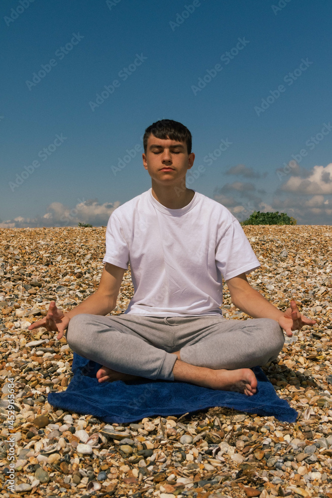 A Caucasian teenage boy meditating on a stony beach with his hands in the Shuni position